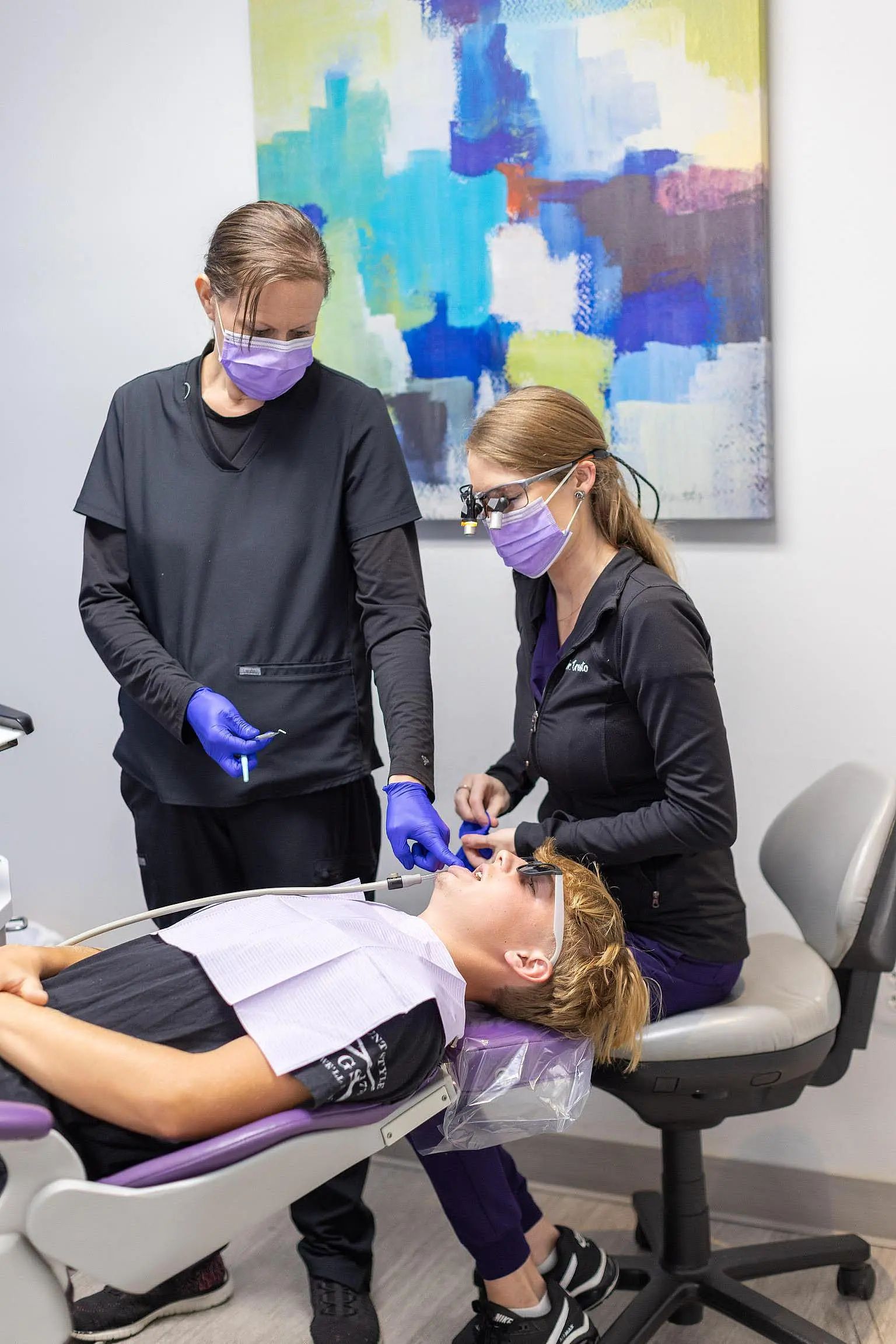 Two dental professionals wearing gloves and masks attend to a patient lying in a dental chair in a modern office with abstract art on the wall at Queen City Orthodontics in Liberty Township or Hamilton, OH.