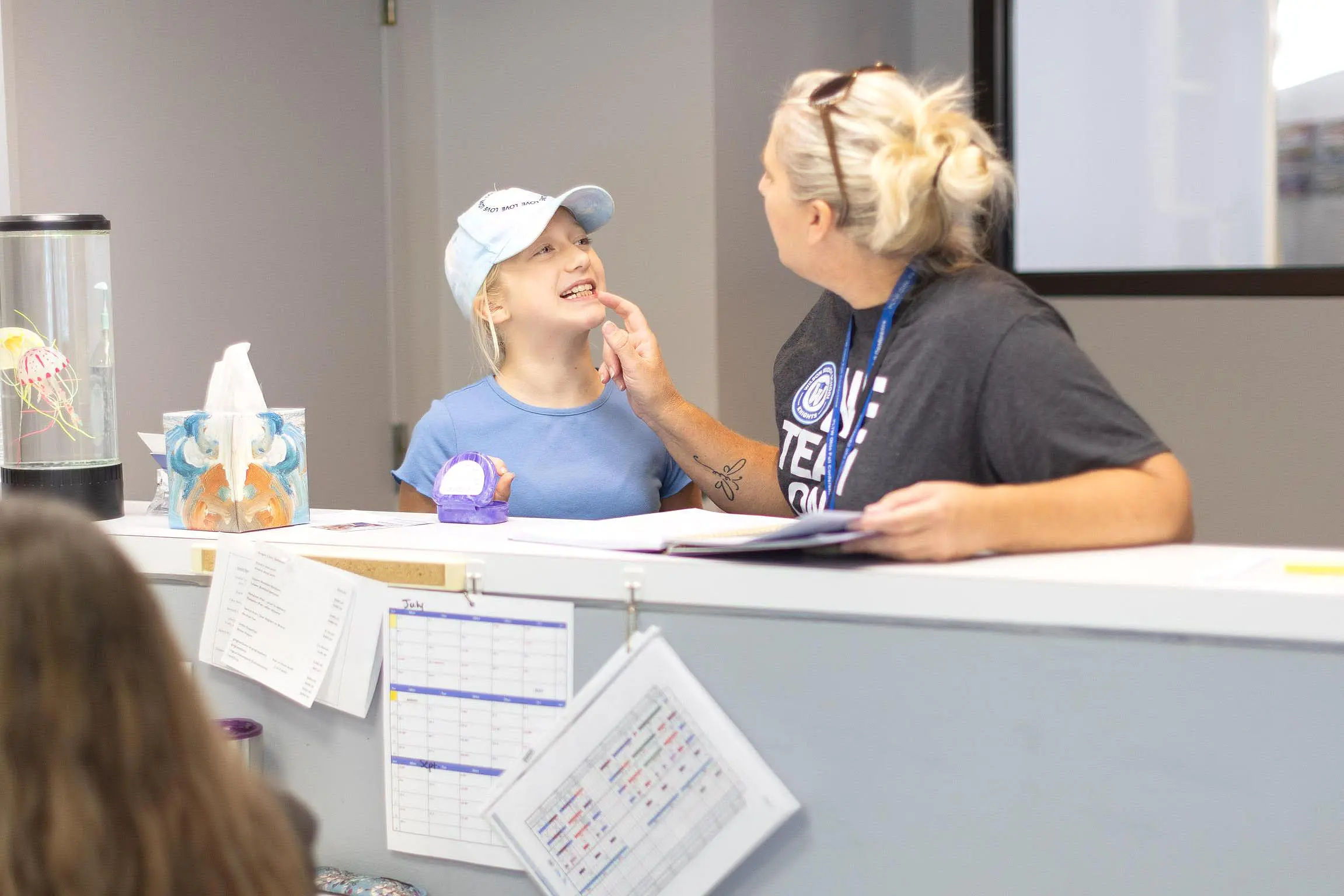 A woman checks a young girl's mouth at a reception desk, both smiling, with papers, a model, and calendars visible on the counter at Queen City Orthodontics in Liberty Township, OH.