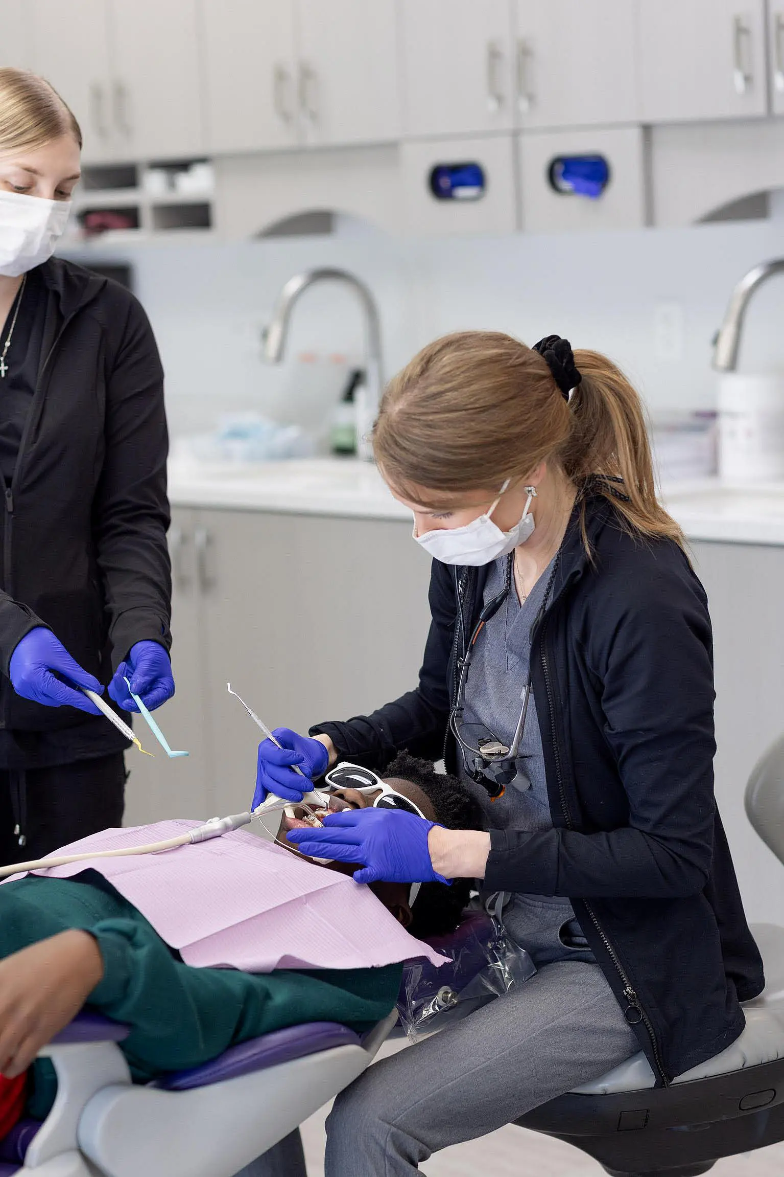 Two dental professionals, wearing masks and gloves, perform a dental procedure on a patient lying in a dental chair in a modern clinic at Queen City Orthodontics in Liberty Township, OH.