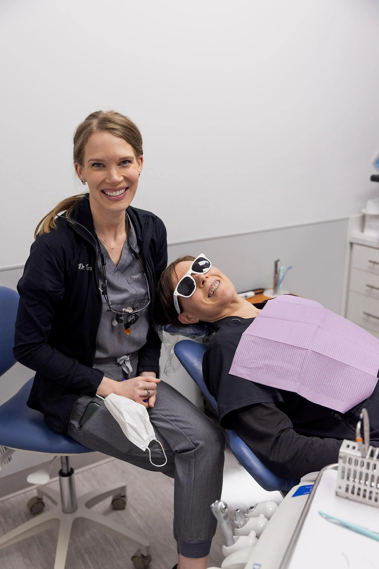 A dentist sits smiling next to a patient reclining in a dental chair, wearing sunglasses and a dental bib in a modern clinic at Queen City Orthodontics in Hamilton, OH.