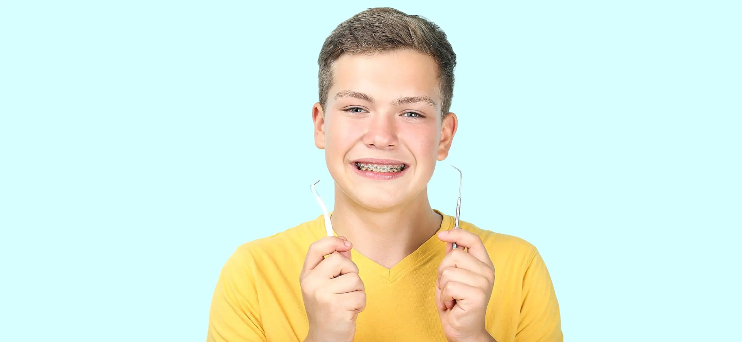A smiling teenage boy in Liberty Township, OH, shows off his new braces while holding a dental mirror and explorer. He's wearing a yellow shirt against a light blue background.
