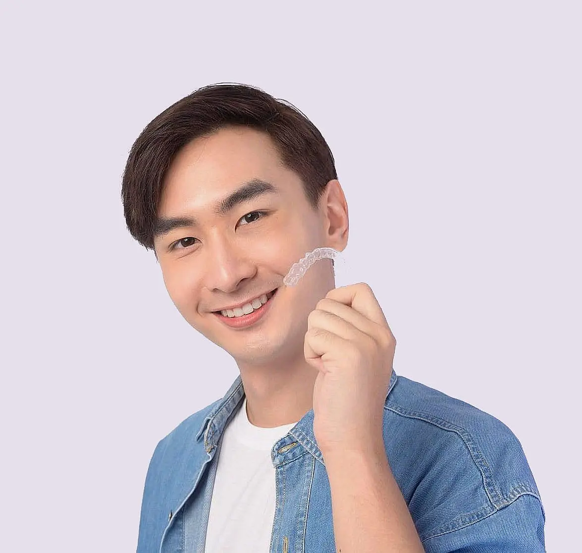 A person with short dark hair, wearing a denim shirt over a white T-shirt, smiles while holding Invisalign® Clear Aligners against a plain background in Hamilton OH