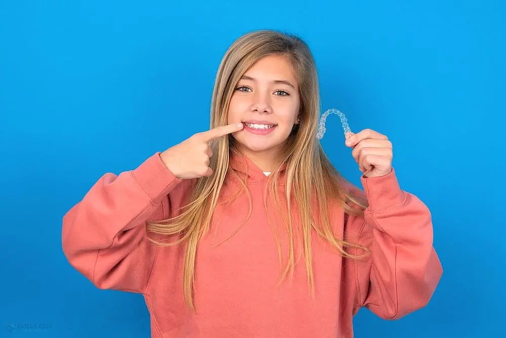 A girl in a pink hoodie stands against a blue background, smiling and pointing at her teeth with one hand while holding a clear dental aligner with the other—showcasing how to fix overbite in Liberty Township or Hamilton, OH.