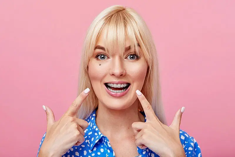 Smiling woman with blonde hair points to her ceramic braces, wearing a blue shirt with white polka dots, standing in front of a pink background in Liberty Township, OH