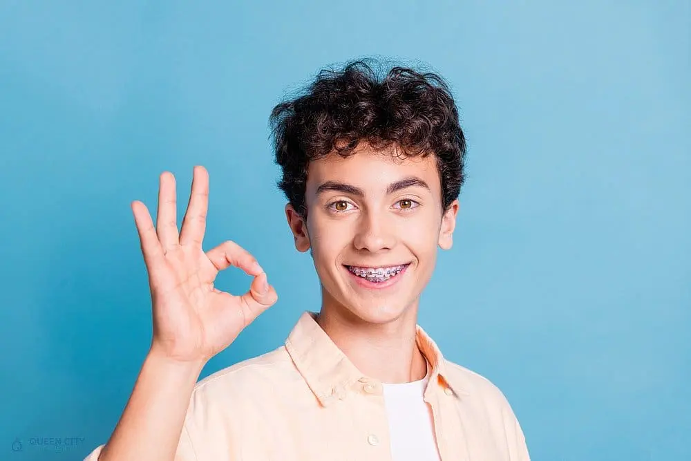 A teen boy with curly hair and braces smiles and gives an "OK" hand gesture against a solid light blue background, representing aligerns vs braces in Liberty Township or Hamilton, OH.