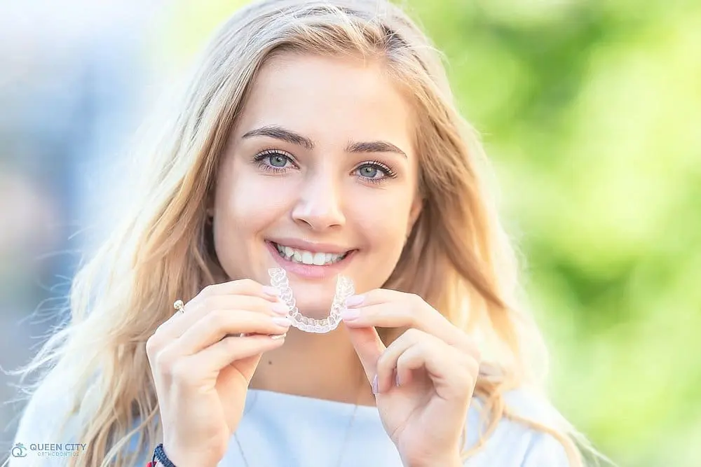 A young woman with long blonde hair smiles outdoors while holding a clear dental aligner in both hands, representing aligners vs braces in Liberty Township or Hamilton, OH.
