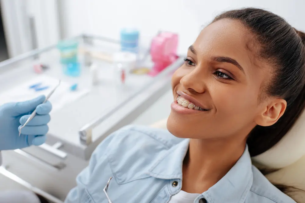 A young woman with braces sits in a dental chair, smiling, while a gloved dentist's hand holds a dental tool nearby in Liberty Township and Hamilton, OH.