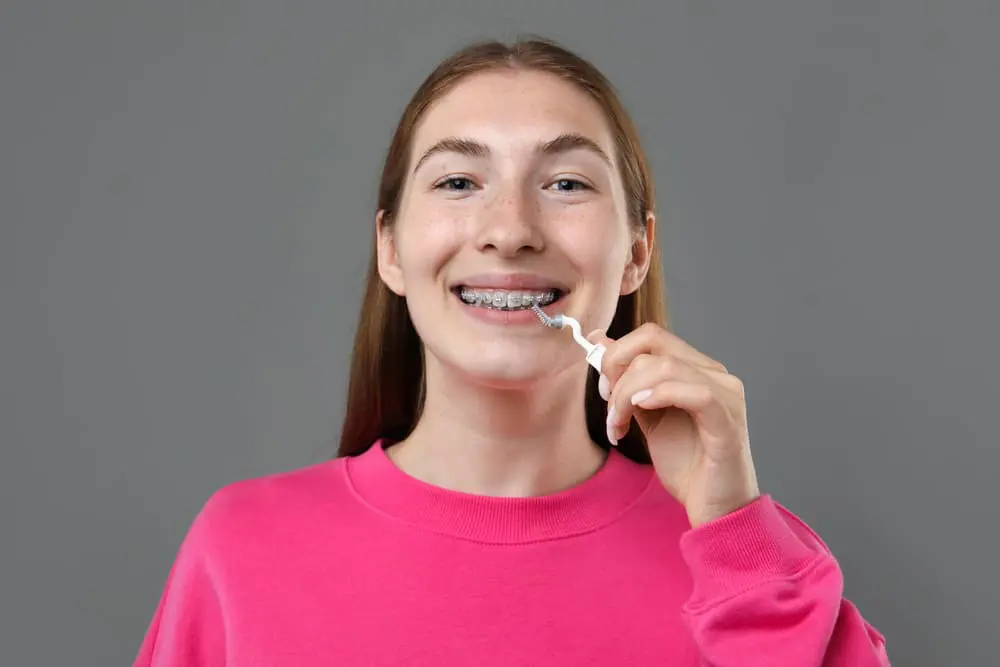 Young person wearing braces uses a dental flosser, smiling at the camera, against a plain gray background in Liberty Township and Hamilton, OH.