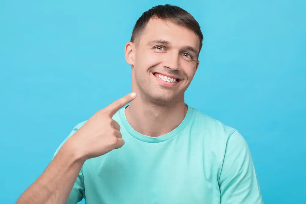 A man wearing a turquoise shirt smiles and points to his dental braces against a solid light blue background in Liberty Township and Hamilton, OH.