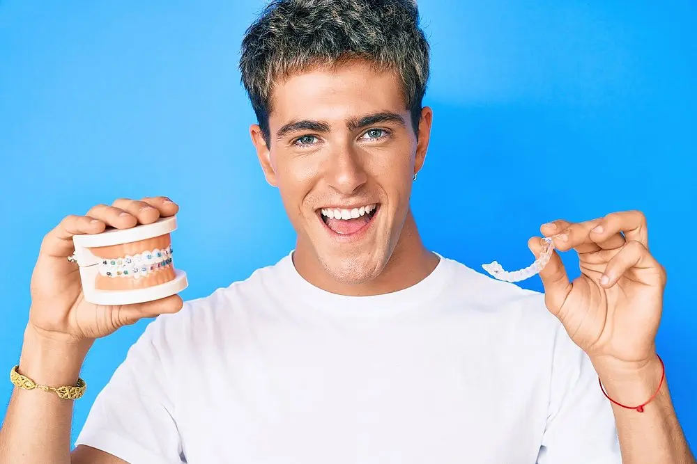aligners vs braces In Liberty Township or Hamilton, OH, Queen City Orthodontics features a person in a white shirt smiling against a blue background while holding a dental model with braces in one hand and a clear aligner in the other.