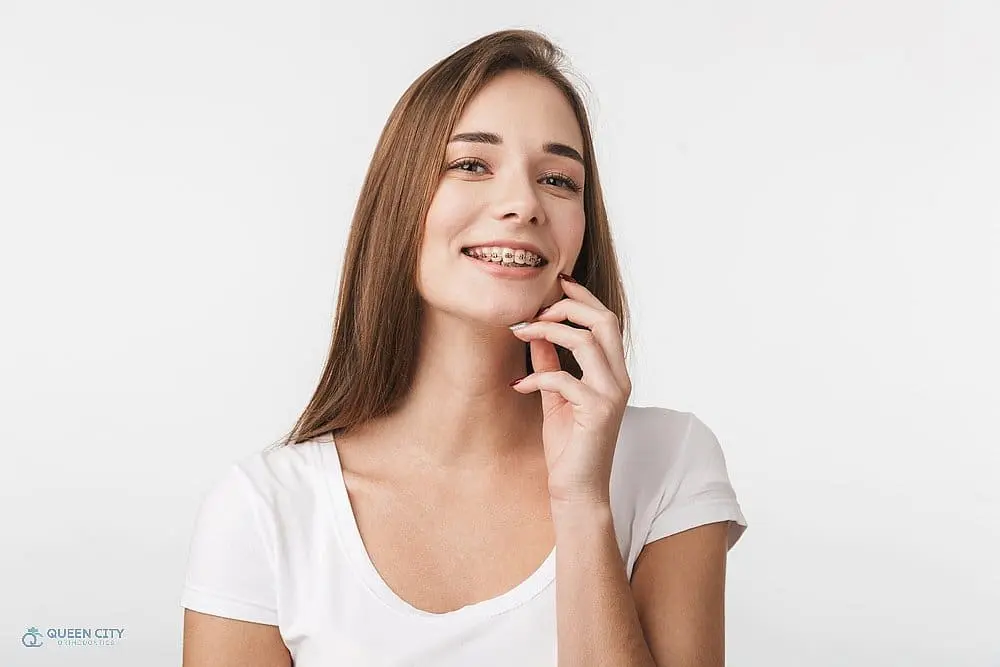 A young woman with straight brown hair, wearing braces and a white shirt, smiles and touches her face wondering about How Long Do Braces Take in Liberty Township and Hamilton, OH against a plain white background—representing patients at Queen City Orthodontics in Liberty Township or Hamilton, OH.