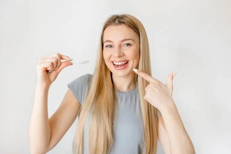 A smiling woman with long blonde hair points to her teeth and holds a clear aligner at Queen City Orthodontics in Liberty Township or Hamilton, OH - can Invisalign fix overbite in Liberty Township and Hamilton, OH.
