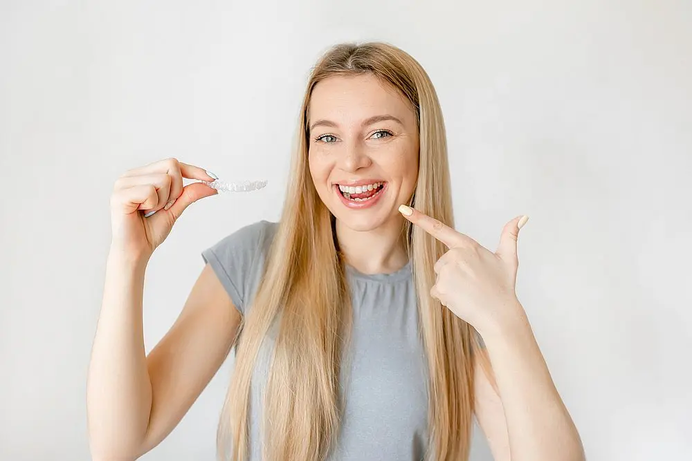 A smiling woman with long blonde hair points to her teeth and holds a clear aligner at Queen City Orthodontics in Liberty Township or Hamilton, OH - can Invisalign fix overbite in Liberty Township and Hamilton, OH.