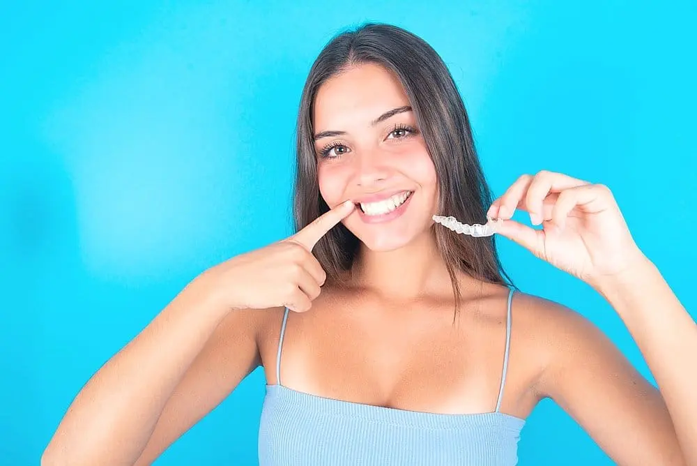 Smiling woman in light blue tank points to her teeth, holding a clear aligner for Queen City Orthodontics in Liberty Township or Hamilton, OH - can Invisalign fix overbite in Liberty Township and Hamilton, OH.
