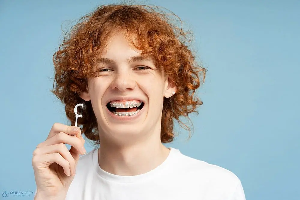 A smiling person with curly red hair and braces holds a dental flosser, representing Queen City Orthodontics in Liberty Township or Hamilton, OH, against a plain blue background - How to Floss with Braces in Liberty Township and Hamilton, OH. 