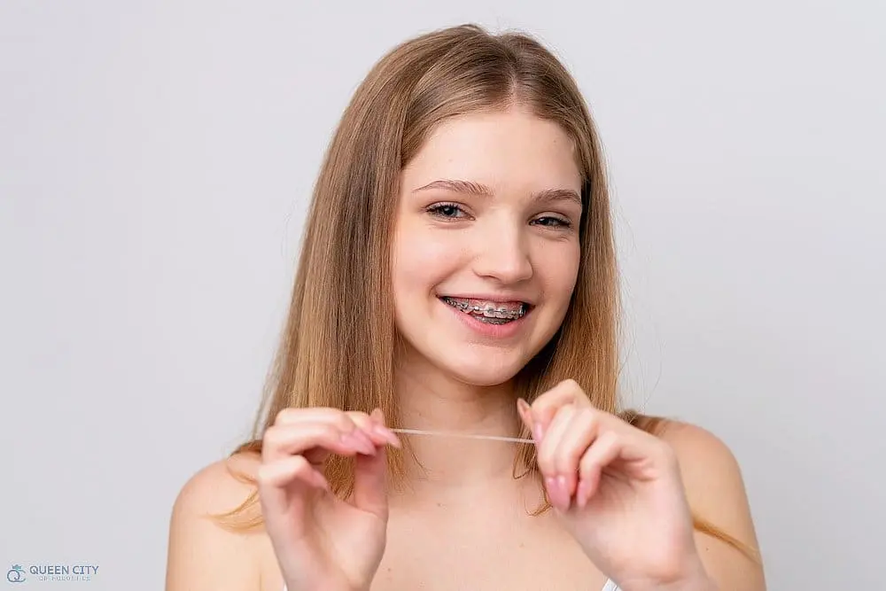 Smiling teen girl with straight, light brown hair and braces holds dental floss, promoting Queen City Orthodontics in Liberty Township or Hamilton, OH - How to Floss with Braces in Liberty Township and Hamilton, OH. 