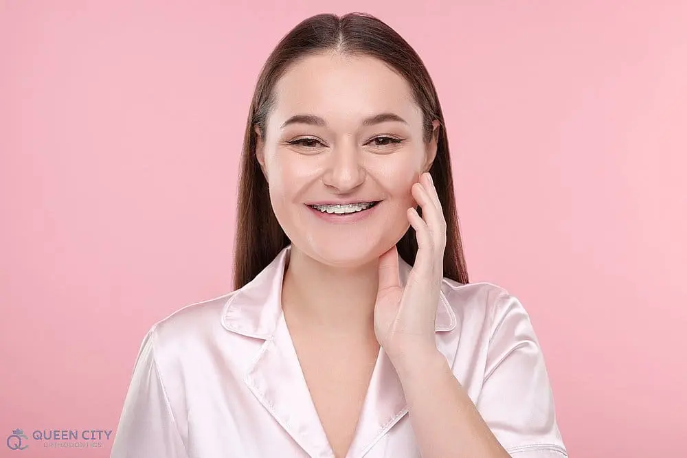 A woman with straight brown hair and braces smiles at Queen City Orthodontics in Liberty Township or Hamilton, OH. Pink background - How to Get Braces Glue Off Teeth in Liberty Township and Hamilton, OH
