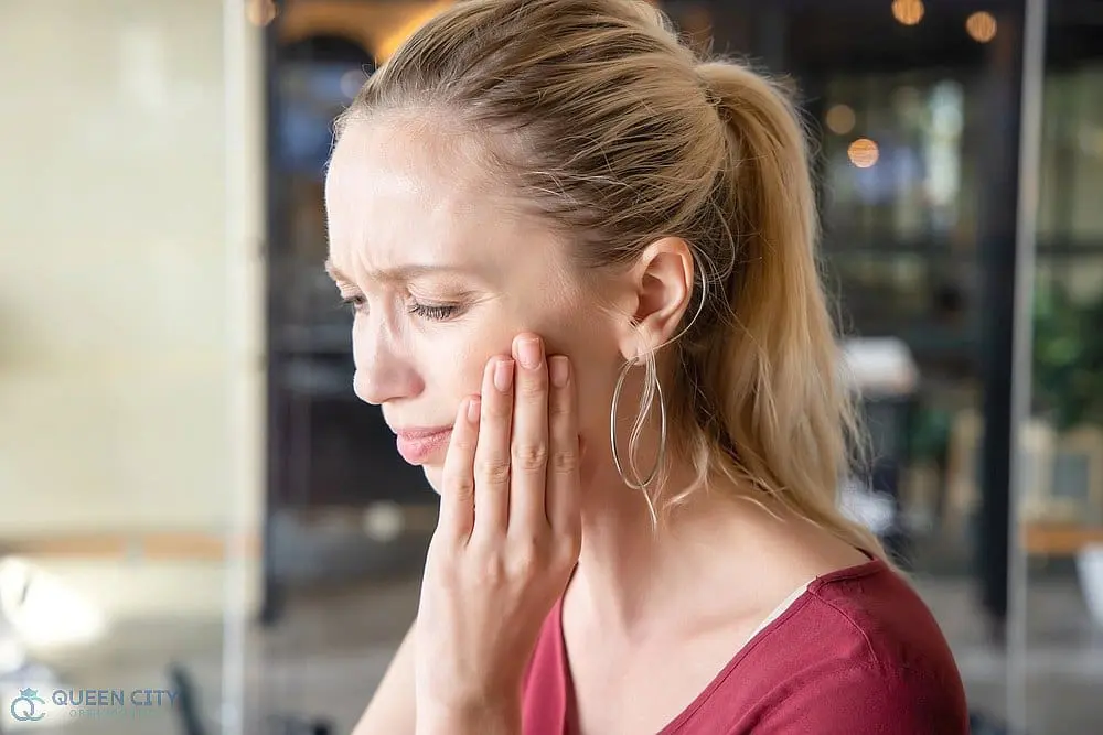 A blonde woman touches her cheek in apparent pain, possibly from a toothache, at Queen City Orthodontics in Liberty Township or Hamilton, OH - TMJ Treatment in Liberty Township and Hamilton, OH