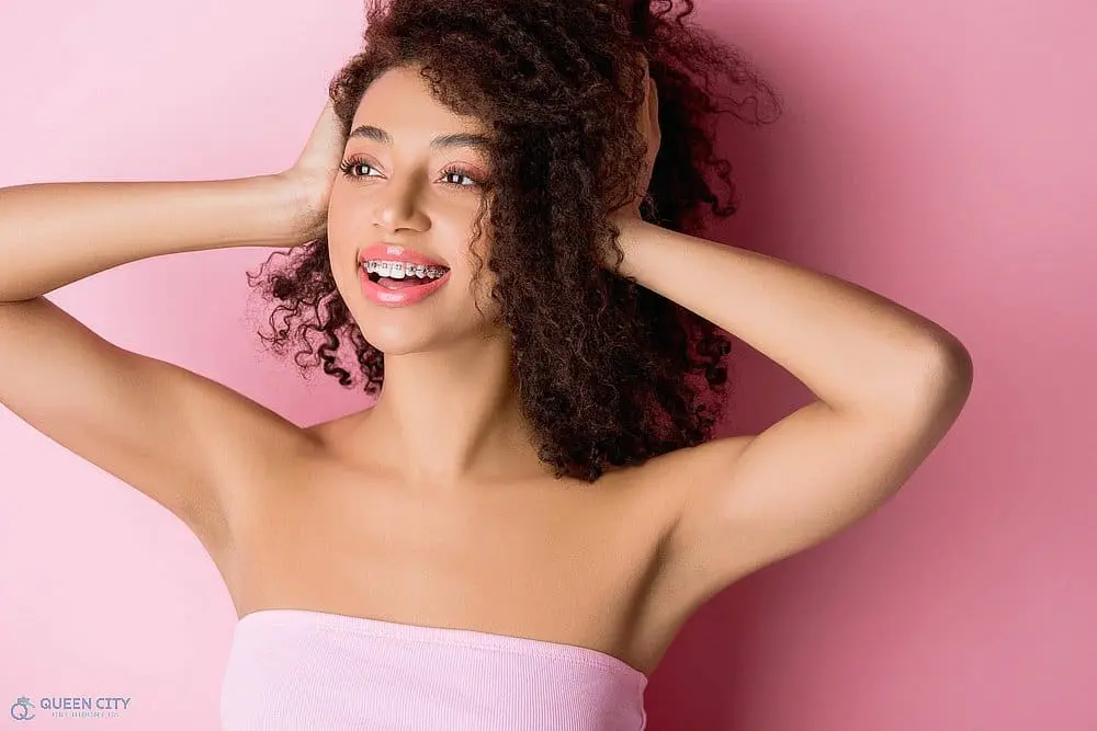 Smiling young woman with curly hair and braces, in a pink top, promotes Queen City Orthodontics in Liberty Township or Hamilton, OH - Clear Braces vs Metal Braces in Liberty Township and Hamilton, OH