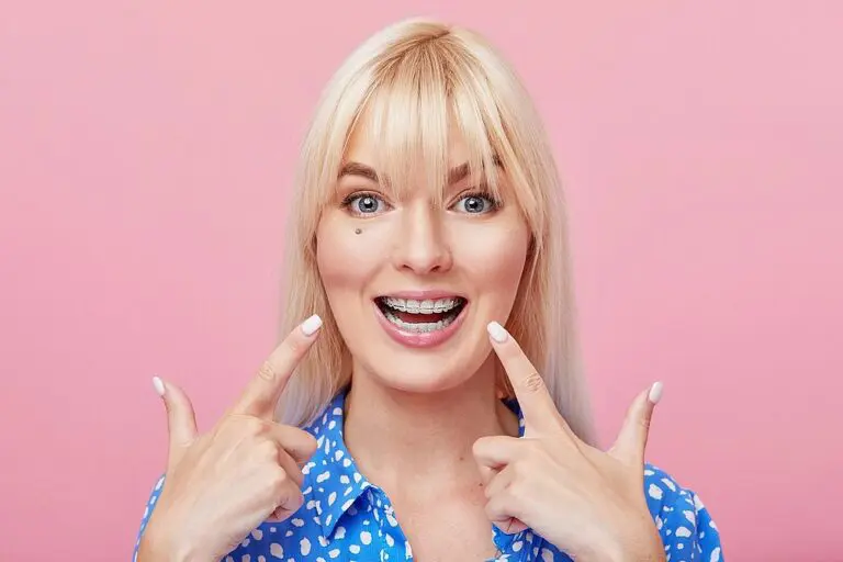 Smiling woman with blonde hair and braces points to her teeth before a pink background at Queen City Orthodontics in Liberty Township or Hamilton, OH - Clear Braces vs Metal Braces in Liberty Township and Hamilton, OH
