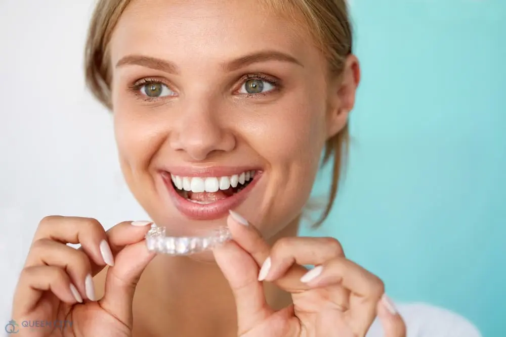 A smiling woman holds a clear dental aligner near her mouth; Queen City Orthodontics in Liberty Township or Hamilton, OH - How Does Invisalign Work in Liberty Township and Hamilton, OH