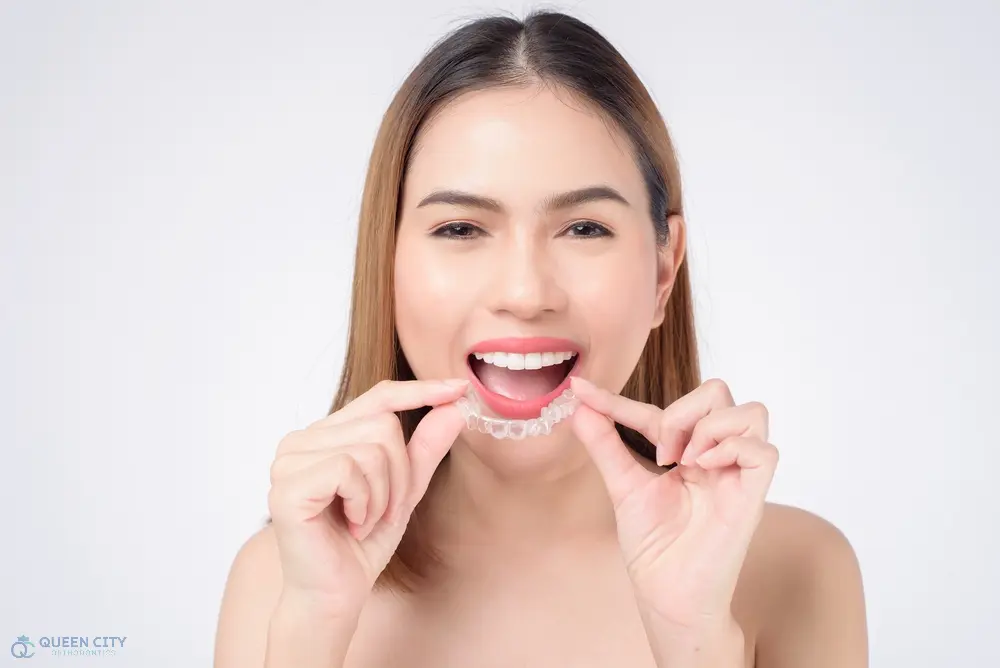 Smiling woman places a clear dental aligner on her teeth for Queen City Orthodontics in Liberty Township or Hamilton, OH - How Long Does Invisalign Take in Liberty Township and Hamilton, OH 