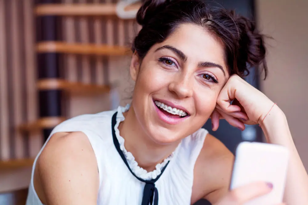 A young woman with braces at Queen City Orthodontics in Liberty Township or Hamilton, OH smiles indoors, holding her smartphone.