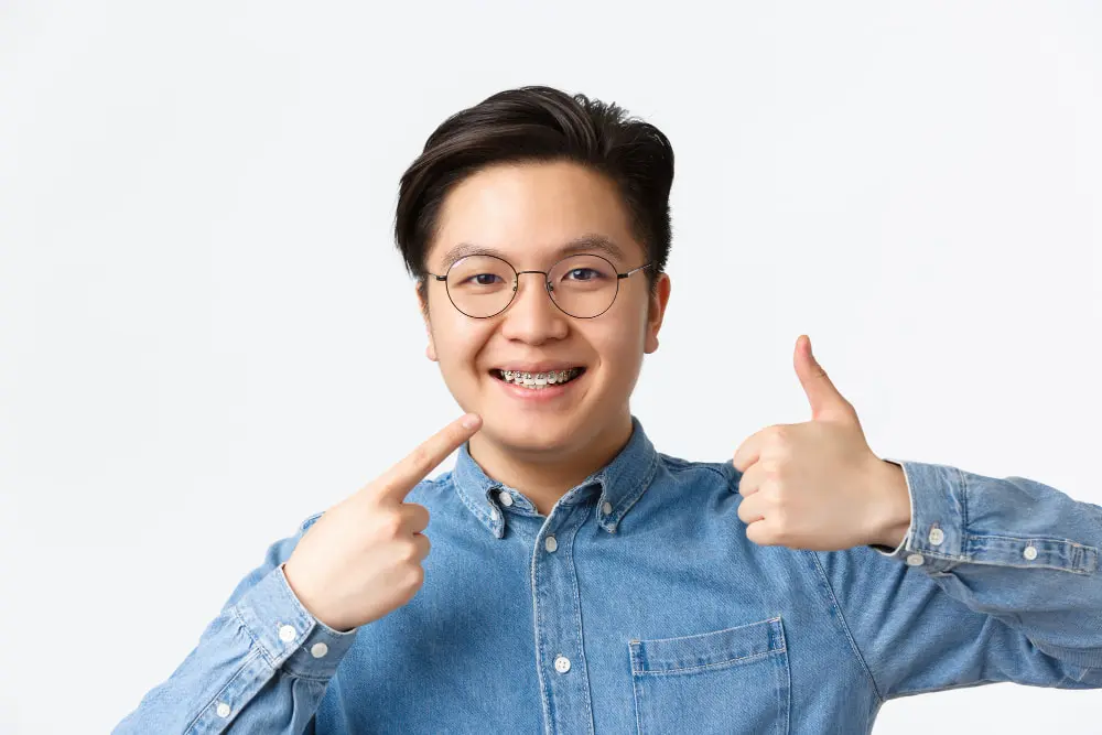 Smiling young person with braces points to their teeth and gives a thumbs up for Queen City Orthodontics in Liberty Township or Hamilton, OH.