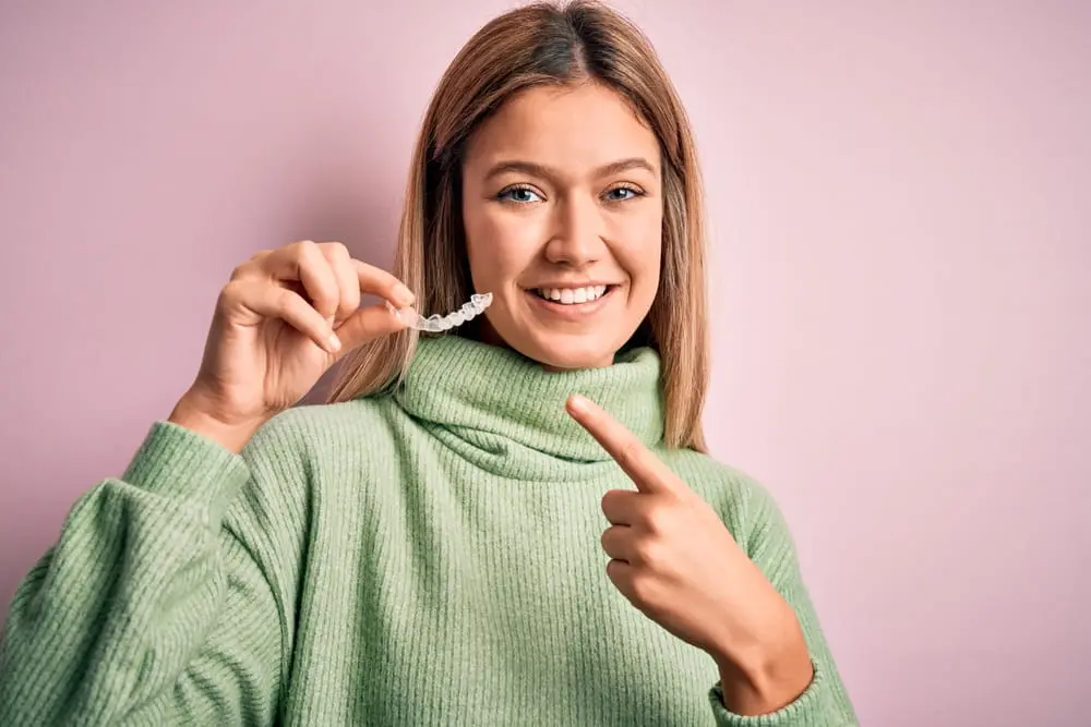 A person in a green sweater smiles while holding and pointing to an Invisalign clear dental aligner against a pink background in Liberty Township, OH