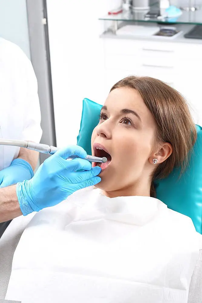 A dentist wearing blue gloves examines a patient's teeth using dental instruments. The patient is seated in a dental chair with a white bib in Liberty Township and Hamilton, OH.