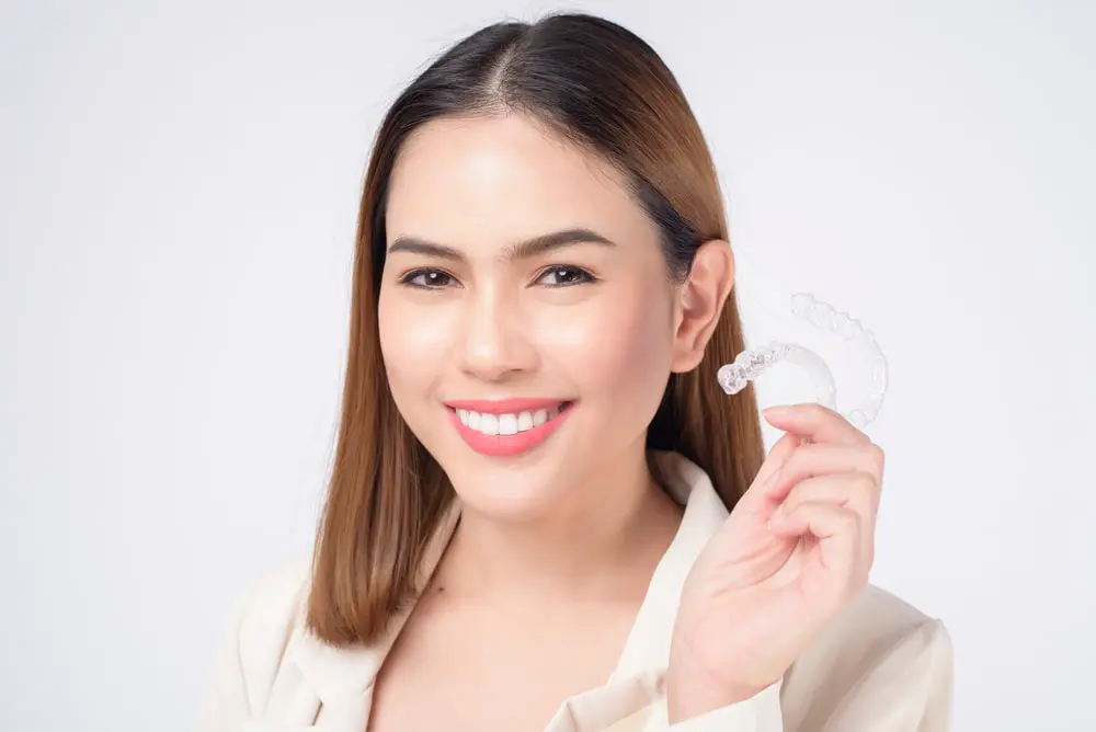A woman smiles while holding Invisalign® Clear Aligners in her right hand, set against a plain white background in Liberty Township OH